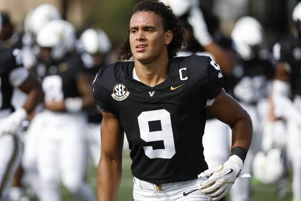 Football player in black jersey with number 9 and captain's C on his chest, on the field with teammates in the background.